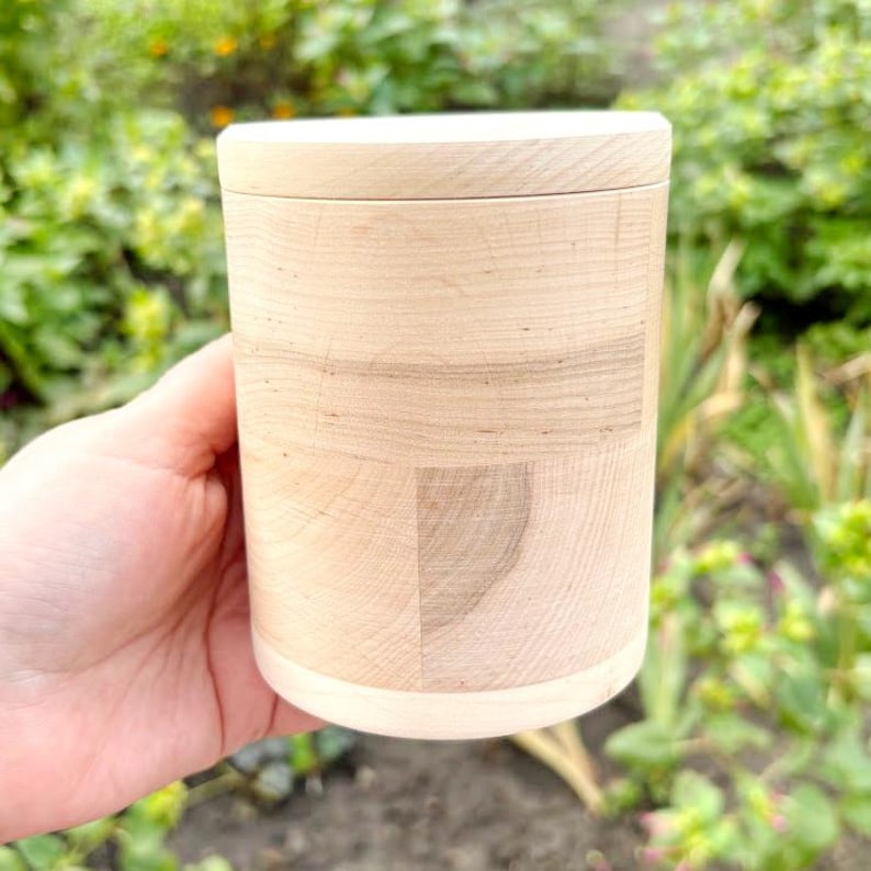 K&ouml;nnte beinhalten: Ein hellfarbener Holzbeh&auml;lter mit Deckel. Der zylindrische Beh&auml;lter besteht aus hellfarbenem Holz mit sichtbaren Maserungsmustern. Der Deckel passt fest auf die Oberseite. Der Beh&auml;lter wird in einer Hand vor einem verschwommenen gr&uuml;nen Hintergrund gehalten.