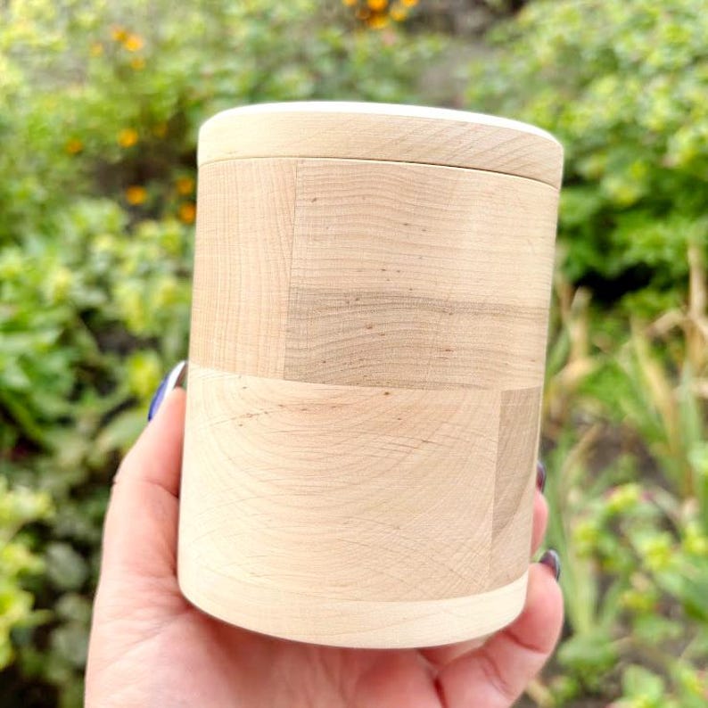 K&ouml;nnte beinhalten: Eine zylindrische Holzkiste mit Deckel. Die Kiste besteht aus hellfarbenem Holz mit einem nat&uuml;rlichen Maserungsmuster. Der Deckel hat einen helleren Wei&szlig;ton. Die Kiste wird in einer Hand vor einem verschwommenen gr&uuml;nen Hintergrund gehalten.
