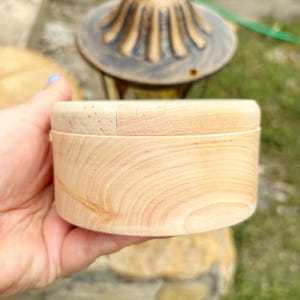Könnte beinhalten: Kleine, runde Holzbox mit Deckel. Die Box besteht aus hellem Holz mit sichtbarer Maserung. Die Box wird in einer Hand gehalten, mit einem verschwommenen Hintergrund aus Grün und einer dekorativen Lampe.
