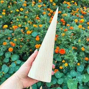 May include: A smooth, light-colored wooden ring display cone with a tapered, pointed top and a wide, circular base. The object features a natural wood grain texture and is designed to hold rings securely on its vertical, conical surface.