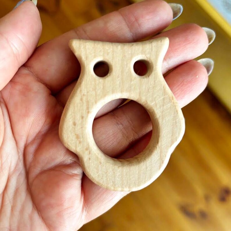 Peut inclure: Un anneau de dentition en bois en forme de hibou. Le jouet est en bois de couleur claire et comporte deux trous circulaires pour les yeux et une grande ouverture circulaire pour la bouche. La t&ecirc;te du hibou a de petites oreilles pointues.