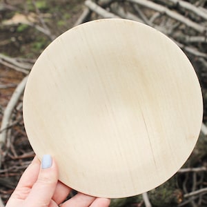May include: A round, light brown wooden plate with a smooth surface. The plate is held in a hand with light blue nail polish.