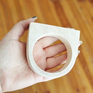 May include: A light-colored wooden ring featuring a thick, flat-topped geometric design with a circular opening. The wood grain is visible across the smooth, pale surface of the accessory, which has a substantial width and a rounded bottom edge.