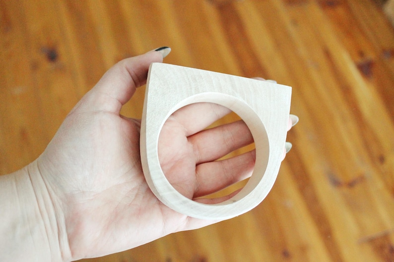 May include: A light-colored wooden ring featuring a thick, flat-topped geometric design with a circular opening. The wood grain is visible across the smooth, pale surface of the accessory, which has a substantial width and a rounded bottom edge.