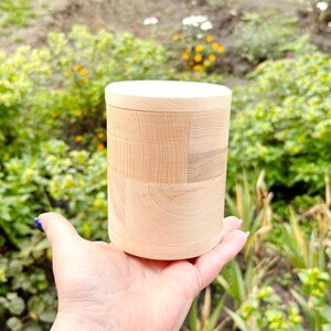 K&ouml;nnte beinhalten: Ein hellfarbiger Holzbeh&auml;lter mit Deckel. Der Beh&auml;lter ist zylindrisch und scheint aus mehreren Holzst&uuml;cken gefertigt zu sein. Der Deckel ist aus massivem, hellfarbigem Holz. Der Beh&auml;lter wird in einer Hand gehalten, mit einem verschwommenen gr&uuml;nen Hintergrund.