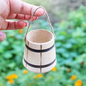 Könnte beinhalten: Ein kleiner Holz-Eimer mit einem braunen Seilhenkel. Der Eimer hat schwarze Bänder um die Ober- und Unterseite.