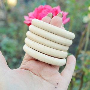 May include: A stack of five natural wood rings, each with a smooth, rounded surface. The rings are held in a hand, with the tips of the fingers visible.
