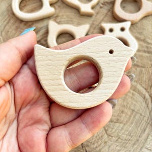 Peut inclure: Une collection de jouets de dentition en bois pour b&eacute;b&eacute;s, en forme d'animaux, dont un oiseau, un &eacute;cureuil et un hibou. Le jouet de dentition en forme d'oiseau est au centre, tenu dans une main, et comporte une ouverture circulaire pour une prise facile.