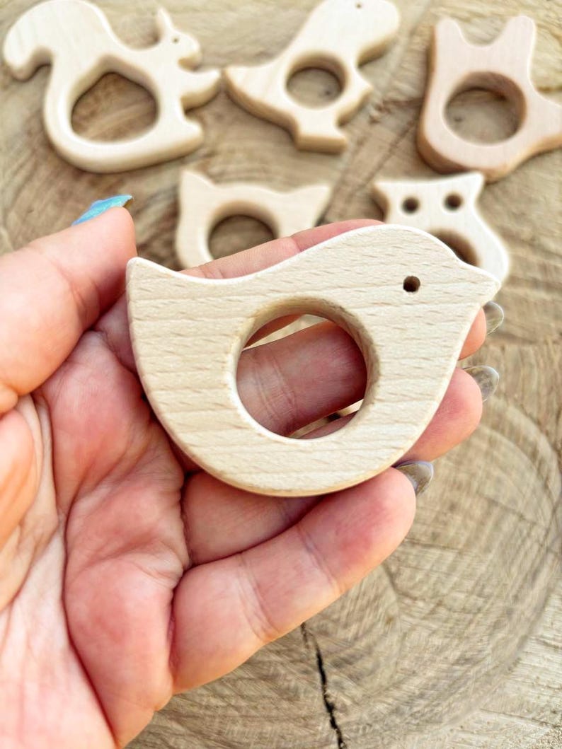 Peut inclure: Une collection de jouets de dentition en bois pour b&eacute;b&eacute;s, en forme d'animaux, dont un oiseau, un &eacute;cureuil et un hibou. Le jouet de dentition en forme d'oiseau est au centre, tenu dans une main, et comporte une ouverture circulaire pour une prise facile.