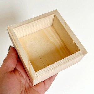 May include: A small, square, unfinished wooden box. The box is constructed from light-colored wood and has a simple, open design. The box is held in a hand, showcasing its size and shape. Ideal for small storage or craft projects.