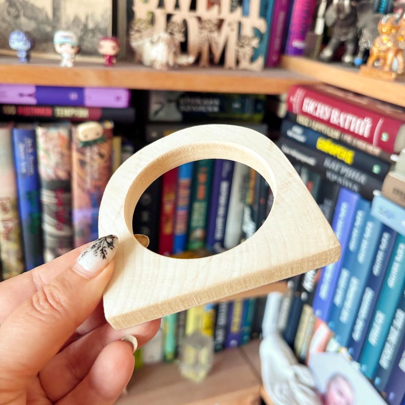 May include: A light-colored, smooth wooden napkin ring featuring a rounded top arch and a flat base. The object has a circular cutout in the center and a natural wood grain finish, showcasing a simple, geometric, and functional design.