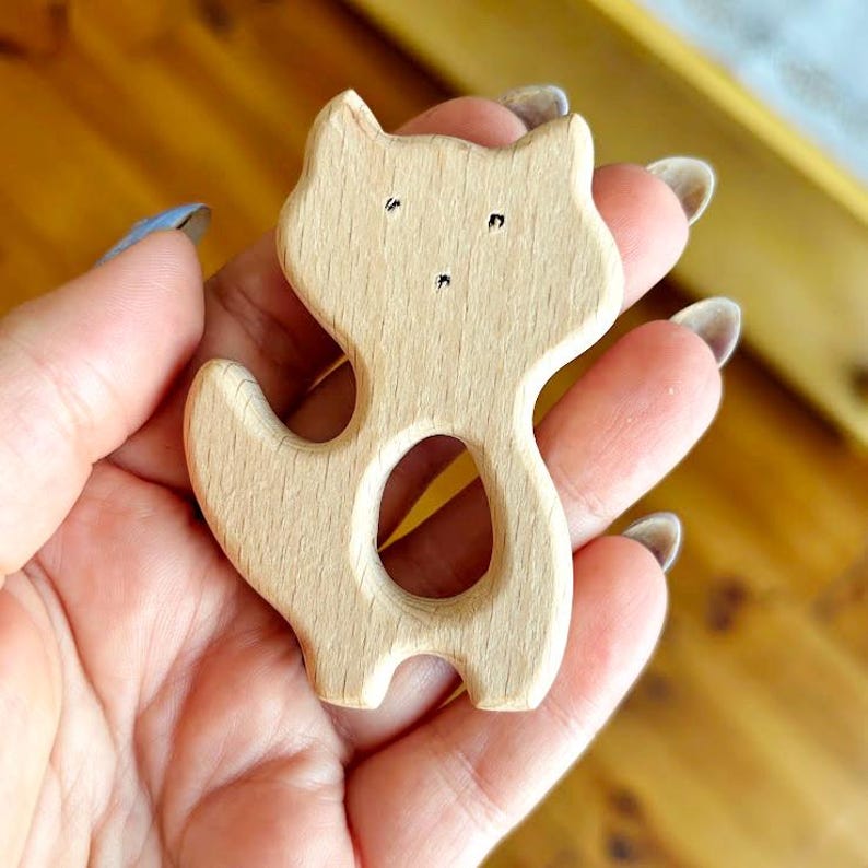 Peut inclure: Un jouet en forme de renard en bois avec une finition en bois naturel. Le renard a un design simple avec de petits points noirs pour les yeux et un nez. Le jouet est tenu dans une main, mettant en valeur sa taille et sa forme. Le renard a une d&eacute;coupe au milieu.