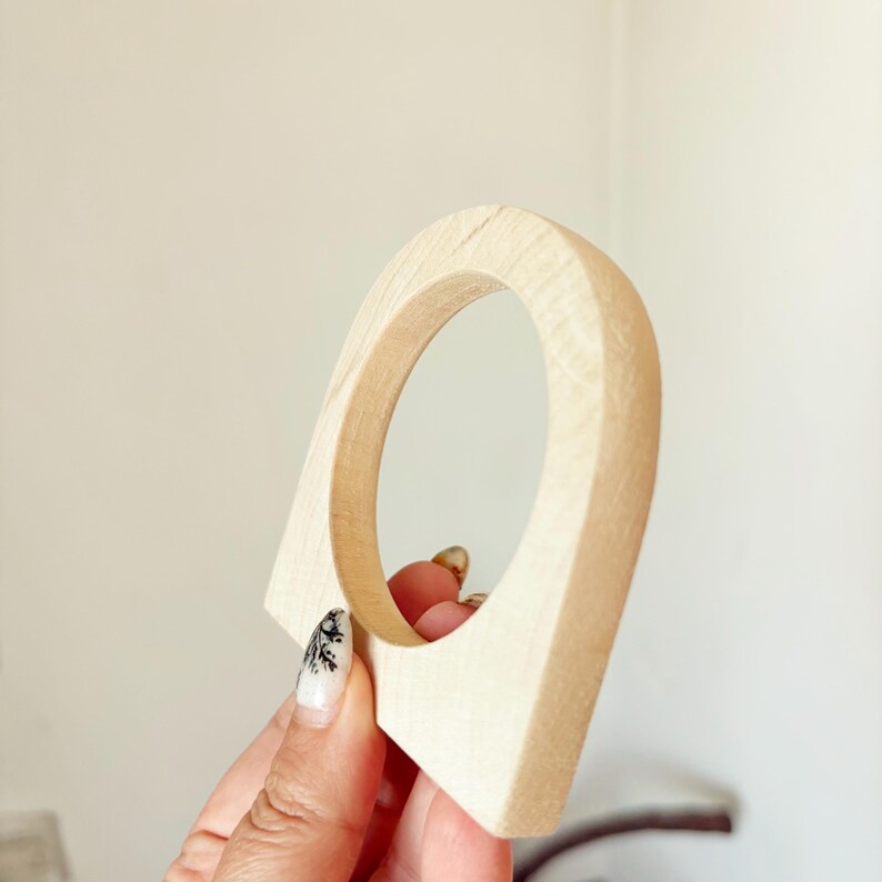 May include: A light-colored, unfinished wooden arch-shaped object with a circular cutout in the center. The piece features a smooth, natural wood grain texture and a flat base, showcasing a simple, geometric form with clean, rounded edges.