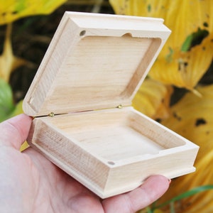 Può includere: Una piccola scatola di legno con un coperchio incernierato, aperta per rivelare un interno vuoto. La scatola è realizzata in legno chiaro e ha una finitura naturale.