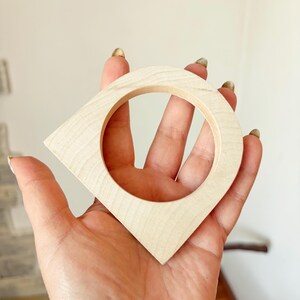 May include: A light-colored wooden object with a smooth, natural grain finish, featuring a circular center cutout and a pointed, triangular shape at the bottom, resembling a map location pin or a stylized geometric pendant or decorative component.