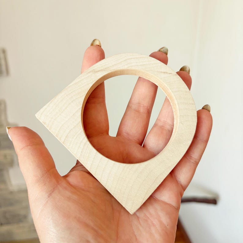 May include: A light-colored wooden object with a smooth, natural grain finish, featuring a circular center cutout and a pointed, triangular shape at the bottom, resembling a map location pin or a stylized geometric pendant or decorative component.