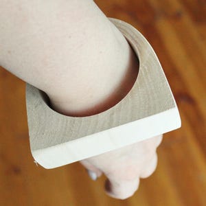 May include: A thick, square-shaped wooden bangle bracelet with a natural light brown wood grain finish on the top and sides, featuring a circular opening in the center for the wrist, showcasing a smooth, solid construction and geometric design.