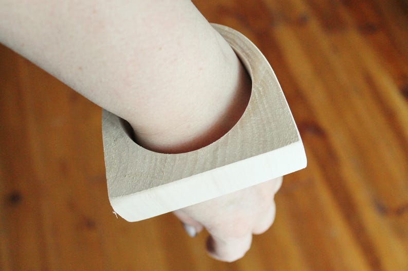 May include: A thick, square-shaped wooden bangle bracelet with a natural light brown wood grain finish on the top and sides, featuring a circular opening in the center for the wrist, showcasing a smooth, solid construction and geometric design.