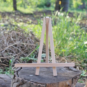 Può includere: Un piccolo cavalletto in legno non rifinito con un design a tre gambe. Il cavalletto è realizzato in legno chiaro e si trova all'aperto su un ceppo d'albero, con uno sfondo verde sfocato di fogliame.