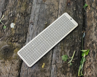Peut inclure: Un marque-page en bois brun clair avec une grille de petits trous. Le marque-page est posé sur une surface en bois vieilli.