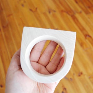 May include: A light-colored wooden ring with a smooth, matte finish and a distinct geometric shape featuring a flat top edge and a circular center opening, showcasing the natural grain patterns of the wood material throughout the surface.
