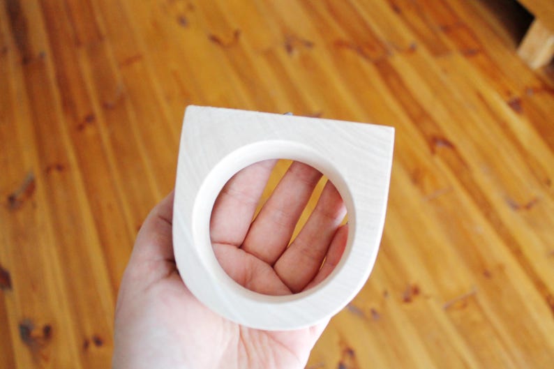 May include: A light-colored wooden ring with a smooth, matte finish and a distinct geometric shape featuring a flat top edge and a circular center opening, showcasing the natural grain patterns of the wood material throughout the surface.