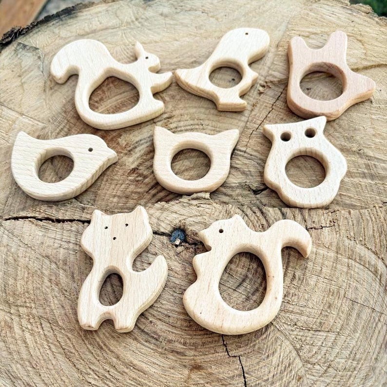 Peut inclure: Ensemble de jouets de dentition en bois en forme d'animaux, dont un &eacute;cureuil, un dinosaure, un oiseau, un chat, un hibou et un renard. Les jouets sont en bois clair et ont une finition lisse. Ils sont dispos&eacute;s sur une surface en bois.