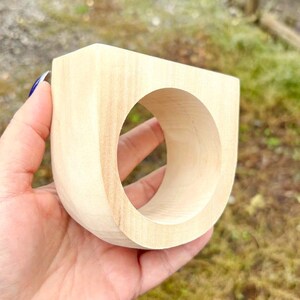 May include: A light-colored, unfinished wooden ring with a thick, U-shaped band and a large circular opening. The wood grain is visible across the smooth, matte surface of the object, which features a flat top edge and rounded sides.