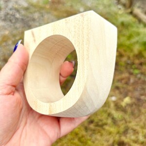 May include: A light-colored, unfinished wooden napkin ring with a smooth, curved exterior and a circular center opening. The natural wood grain is visible across the surface, showcasing the raw texture and pale beige tones of the material.