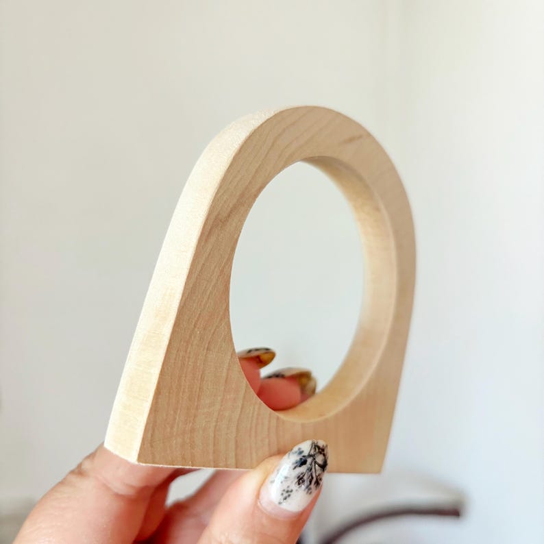 May include: A light-colored wooden object with a smooth, arched top and a circular cutout in the center. The piece features a natural wood grain pattern with a flat base and curved sides, showcasing a solid construction and a light beige finish.