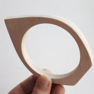 May include: A simple wooden bangle with a rounded, oval shape. The bangle is made of light-colored wood and has a smooth finish.