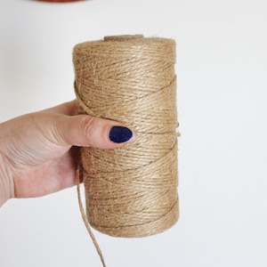 May include: A roll of natural brown twine, wrapped in a spiral, with a loose end hanging down. The twine is held in a hand with dark blue nail polish.