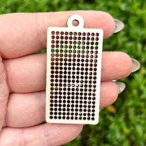 Peut inclure: Un pendentif rectangulaire en bois clair pr&eacute;sentant une grille de petites d&eacute;coupes circulaires r&eacute;guli&egrave;rement espac&eacute;es. Le pendentif a une surface lisse et plate avec un petit trou circulaire au centre sup&eacute;rieur pour la fixation, r&eacute;v&eacute;lant le grain du bois.