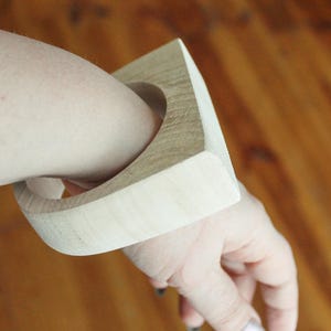 May include: A wide, light-colored wooden bangle bracelet with a square-shaped outer profile and a circular inner opening. The natural wood grain is visible across the smooth, matte surface of the thick, rigid accessory worn on the wrist.
