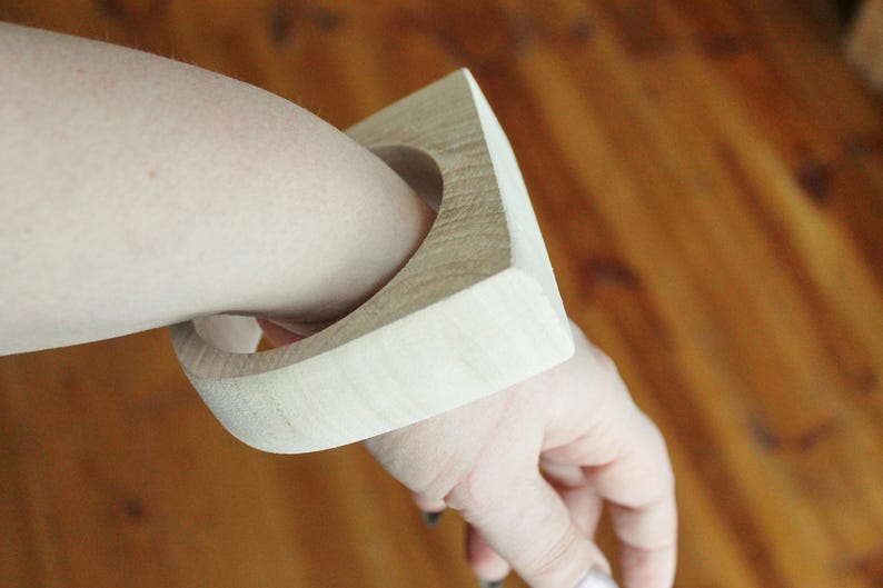 May include: A wide, light-colored wooden bangle bracelet with a square-shaped outer profile and a circular inner opening. The natural wood grain is visible across the smooth, matte surface of the thick, rigid accessory worn on the wrist.