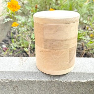 K&ouml;nnte beinhalten: Ein hellfarbener Holzbeh&auml;lter mit Deckel. Der zylindrische K&ouml;rper besteht aus rechteckigen Holzst&uuml;cken, wodurch ein Patchwork-Effekt entsteht. Der Deckel und der Boden sind aus massivem Holz. Der Beh&auml;lter steht vor einem Hintergrund aus gr&uuml;nem Laub und gelben Blumen.