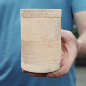 K&ouml;nnte beinhalten: Ein zylindrischer Holzbeh&auml;lter mit Deckel, der von einer Person in der Hand gehalten wird. Der Beh&auml;lter besteht aus hellem Holz mit sichtbaren Maserungsmustern. Das Design ist schlicht und nat&uuml;rlich, was auf einen Aufbewahrungsgegenstand oder ein Dekorationsobjekt hindeutet.