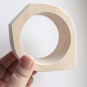 Puede incluir: Un anillo de madera clara con una superficie lisa y redondeada. El anillo se sostiene en una mano, y el interior del anillo es visible.