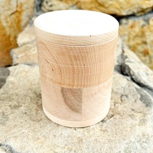 K&ouml;nnte beinhalten: Ein zylindrischer Holzbeh&auml;lter mit hellem Deckel. Der K&ouml;rper des Beh&auml;lters besteht aus hellfarbenem Holz mit sichtbaren Maserungsmustern. Der Beh&auml;lter steht auf einer hellfarbenen Steinoberfl&auml;che.