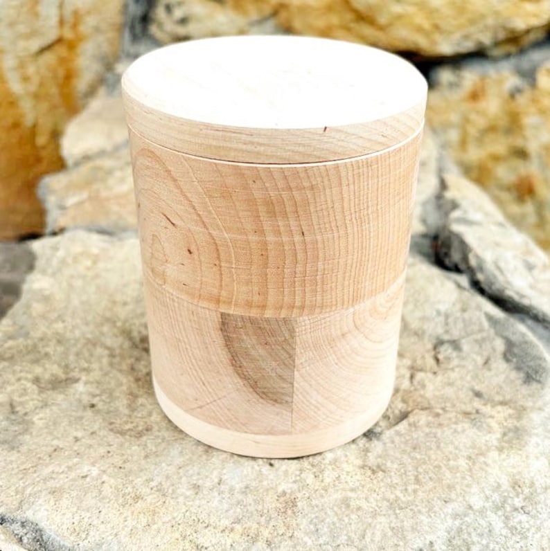 K&ouml;nnte beinhalten: Ein zylindrischer Holzbeh&auml;lter mit hellem Deckel. Der K&ouml;rper des Beh&auml;lters besteht aus hellfarbenem Holz mit sichtbaren Maserungsmustern. Der Beh&auml;lter steht auf einer hellfarbenen Steinoberfl&auml;che.