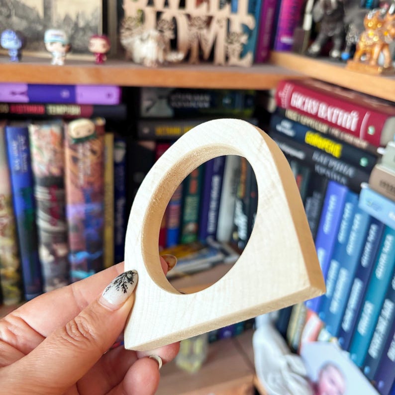 May include: A light-colored, unfinished wooden arch-shaped object with a circular cutout in the center. The piece has a smooth, flat base and a curved top, showcasing the natural grain and texture of the wood in a simple, geometric form.