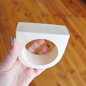 May include: A light-colored, unfinished wooden napkin ring with a square outer profile and a circular center opening. The wood grain is visible on the smooth, matte surface of the object, which features a flat top edge and curved sides.