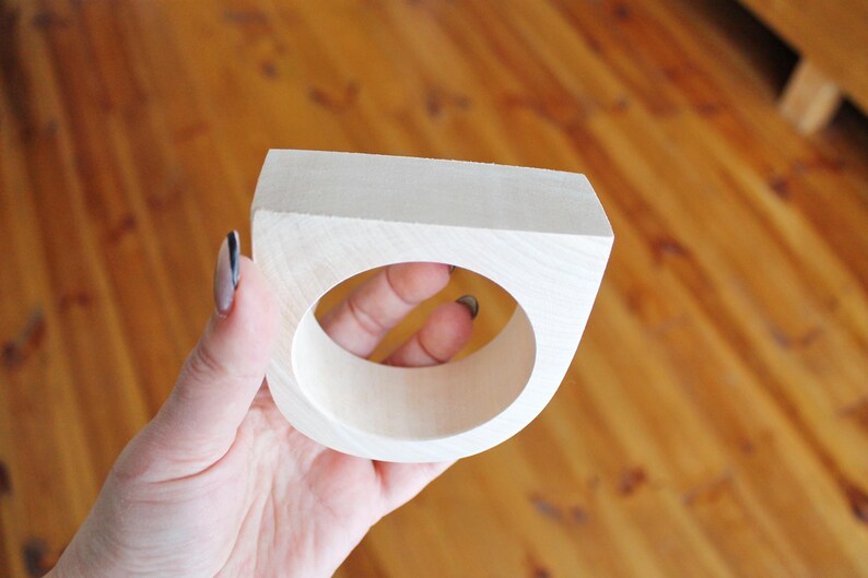 May include: A light-colored, unfinished wooden napkin ring with a square outer profile and a circular center opening. The wood grain is visible on the smooth, matte surface of the object, which features a flat top edge and curved sides.
