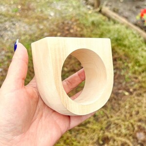 May include: A thick, light-colored wooden ring with a smooth, unfinished surface. The ring features a wide, U-shaped design with a large circular opening in the center, showcasing the natural grain and pale beige tone of the wood material.