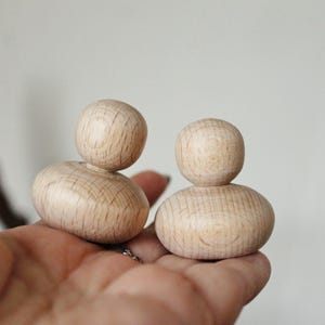 May include: Two wooden stacking dolls, each with a round head and a slightly wider, rounded base. The dolls are made of light-coloured wood and are stacked on top of each other.