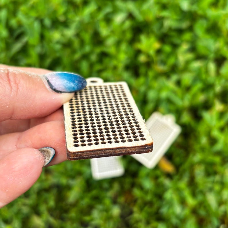 Peut inclure: Un pendentif rectangulaire en bois pr&eacute;sentant une grille de trous d&eacute;coup&eacute;s au laser. La surface est en bois clair avec des bords sombres et br&ucirc;l&eacute;s autour de chaque perforation circulaire. L'objet a une structure plate et un petit trou.