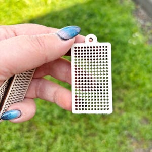 Peut inclure: Un pendentif rectangulaire en bois blanc pr&eacute;sentant une grille de petites d&eacute;coupes circulaires r&eacute;guli&egrave;rement espac&eacute;es et un trou unique en haut pour la suspension. L'objet a une surface plate et un aspect d&eacute;coup&eacute; au laser net.