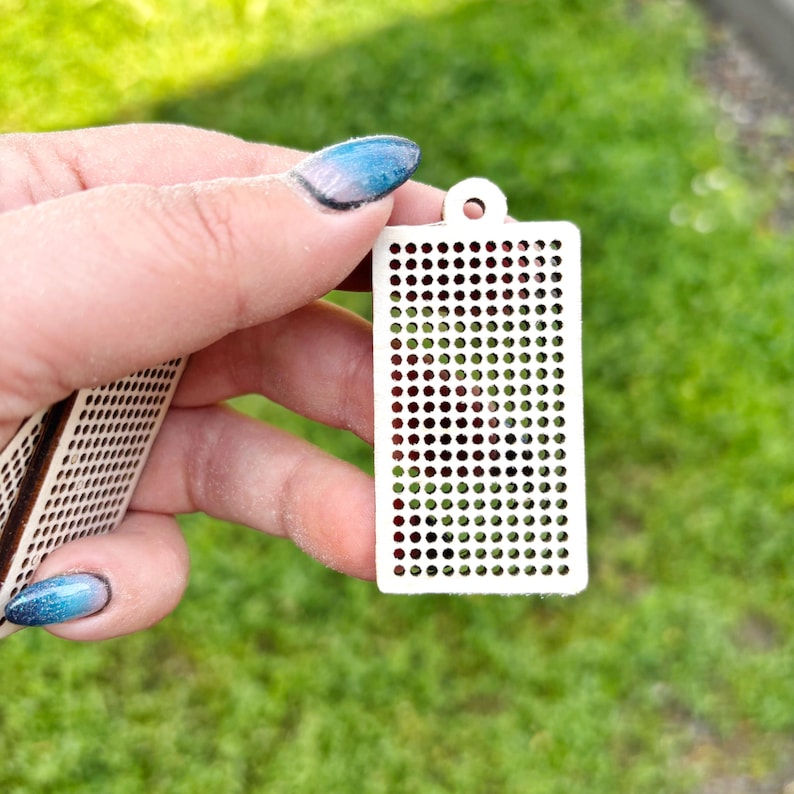 Peut inclure: Un pendentif rectangulaire en bois blanc pr&eacute;sentant une grille de petites d&eacute;coupes circulaires r&eacute;guli&egrave;rement espac&eacute;es et un trou unique en haut pour la suspension. L'objet a une surface plate et un aspect d&eacute;coup&eacute; au laser net.