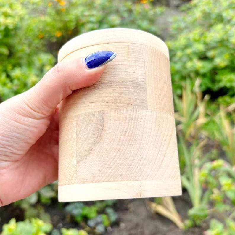 K&ouml;nnte beinhalten: Ein zylindrischer Holzbeh&auml;lter mit Deckel. Der Beh&auml;lter besteht aus hellfarbenem Holz mit sichtbaren Maserungsmustern. Der Deckel ist abgerundet und der K&ouml;rper des Beh&auml;lters ist gerade. Der Beh&auml;lter wird von einer Hand mit einem blauen Fingernagel gehalten.