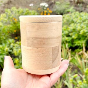 K&ouml;nnte beinhalten: Ein zylindrischer Holzbeh&auml;lter mit Deckel, gehalten in einer Hand. Der Beh&auml;lter besteht aus hellem Holz mit sichtbarer Maserung und einem geometrischen Muster. Der Deckel sitzt b&uuml;ndig oben. Der Hintergrund ist mit gr&uuml;nem Laub verschwommen.
